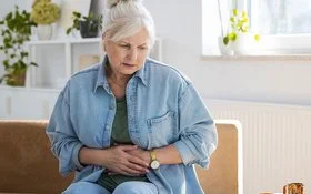 Eine Frau sitzt mit einem ernsten Gesichtsausdruck auf dem Sofa und hält sich die Hände an den Bauch.