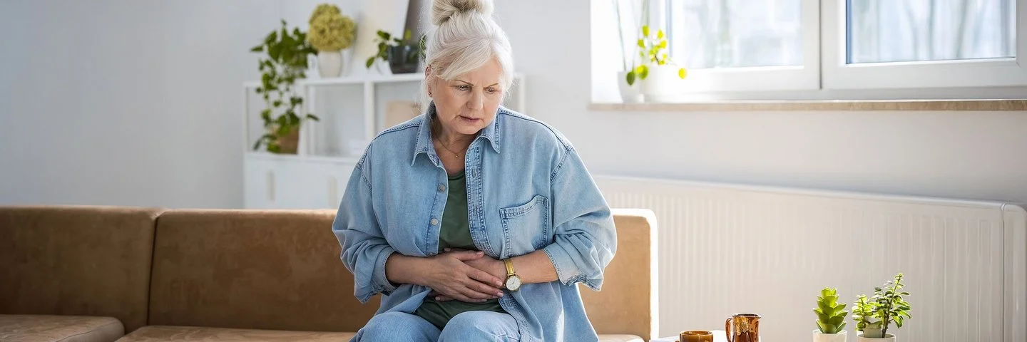 Eine Frau sitzt mit einem ernsten Gesichtsausdruck auf dem Sofa und hält sich die Hände an den Bauch.