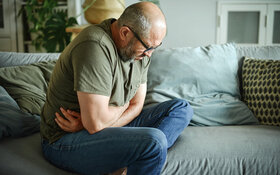 Ein Mann sitzt mit einem Magen-Darm-Infekt schmerzerfüllt auf dem Sofa.