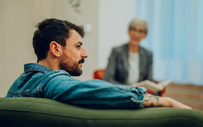Ein junger Mann sitzt auf einem Sofa. Im Hintergrund ist unscharf eine ältere Frau zu erkennen. Die Szene wirkt wie eine Therapiesitzung.