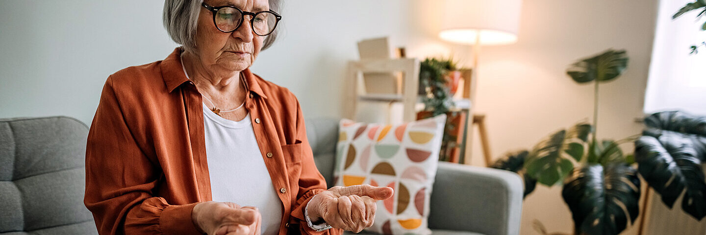 Eine Seniorin sitzt auf der Couch und schaut auf ihre schmerzhaft verkrampften Hände.