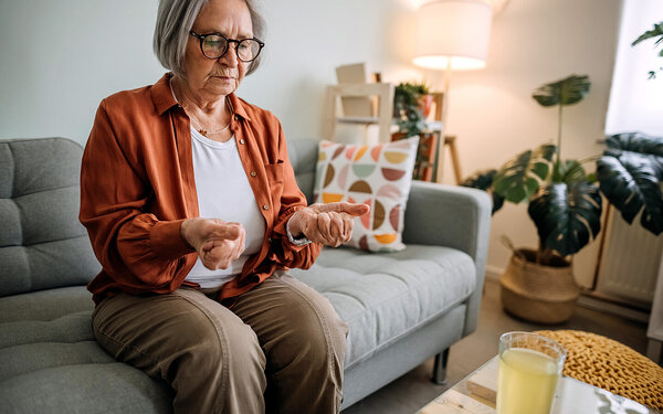 Eine Seniorin sitzt auf der Couch und schaut auf ihre schmerzhaft verkrampften Hände.