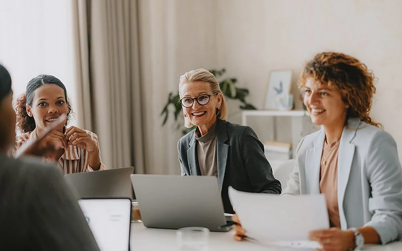 In einem Arbeitsumfeld sitzen vier Frauen mit Laptops an einem Tisch und blicken sich freundlich an.