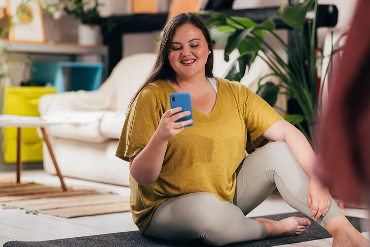 Eine Frau mit Übergewicht sitzt in Sportkleidung auf einer Yogamatte in einem Wohnzimmer. Sie hält ein Smartphone in der Hand und lächelt beim Blick auf den Bildschirm. Im Hintergrund ist ein gemütliches Wohnzimmer mit Pflanzen, einer hellen Couch und verschiedenen Wohnaccessoires zu sehen.