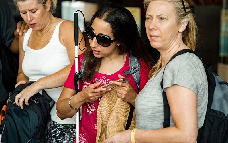 Junge Frau mit Brille und Blindenstock steht in einer Gruppe, mit Smartphone und Strohhut.
