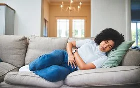 Eine junge Frau liegt mit Bauchkrämpfen auf dem Sofa.