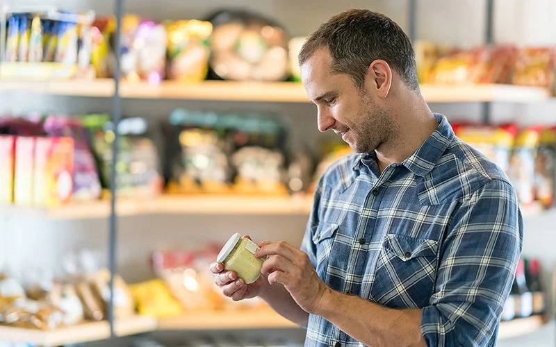 Ein Mann steht in einem Geschäft vor einem Regal mit Produkten. Er liest das Etikett eines Lebensmittelglases.