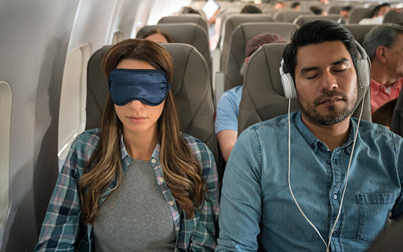 Eine Frau mit Schlafmaske und ein Mann mit Kopfhörern schlafen im Flugzeug.