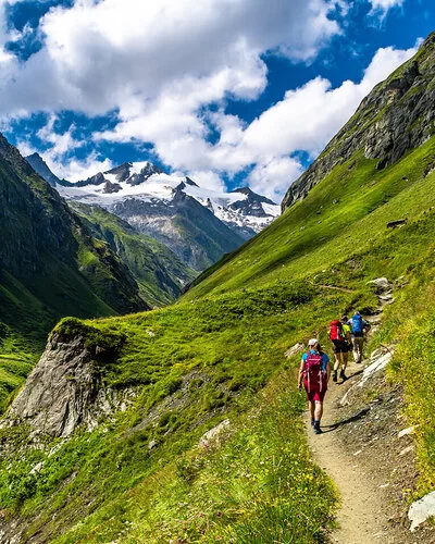 An einem Sommertag macht sich eine dreiköpfige Wandergruppe aus einem Tal auf den Weg zum Gipfel.