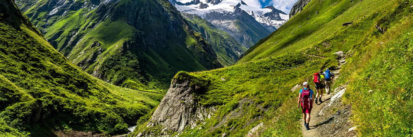 An einem Sommertag macht sich eine dreiköpfige Wandergruppe aus einem Tal auf den Weg zum Gipfel.