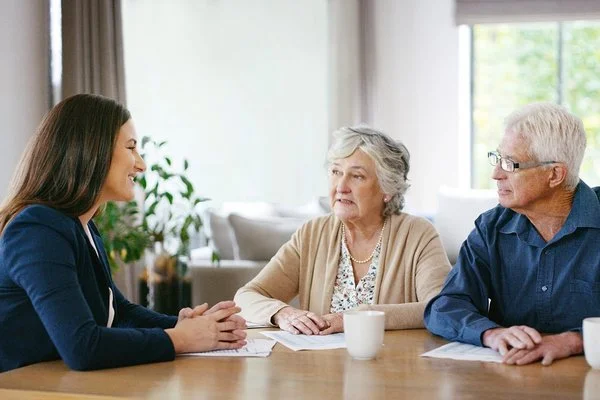 Ein älterer Mann und eine ältere Frau sitzen einer jüngeren Frau gegenüber, die die beiden anlächelt.
