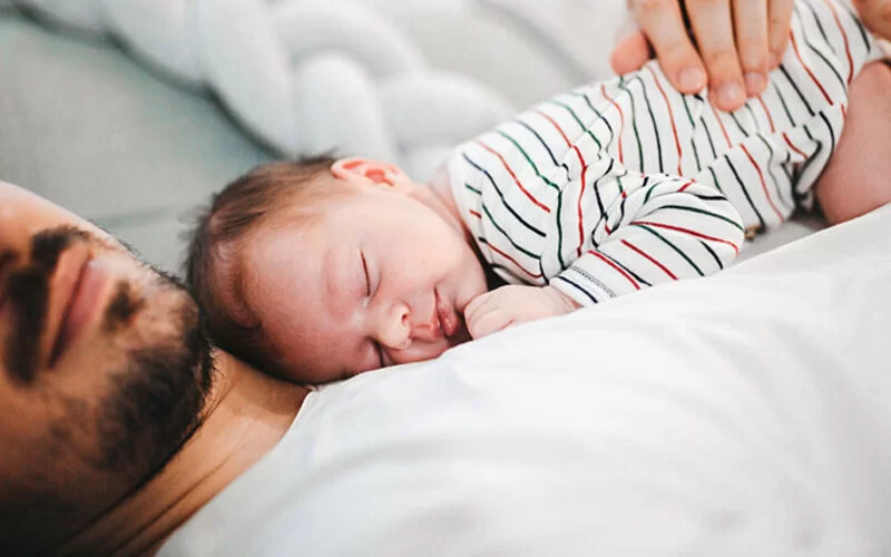 Vater hält sein Baby im Arm, beide schlafen.