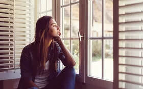 Eine junge Frau sitzt, das Kinn in eine Hand gestützt, an einem Fenster und schaut nach draußen.