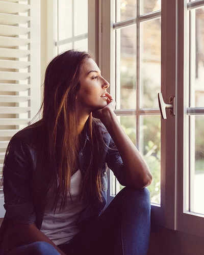 Eine junge Frau sitzt, das Kinn in eine Hand gestützt, an einem Fenster und schaut nach draußen.