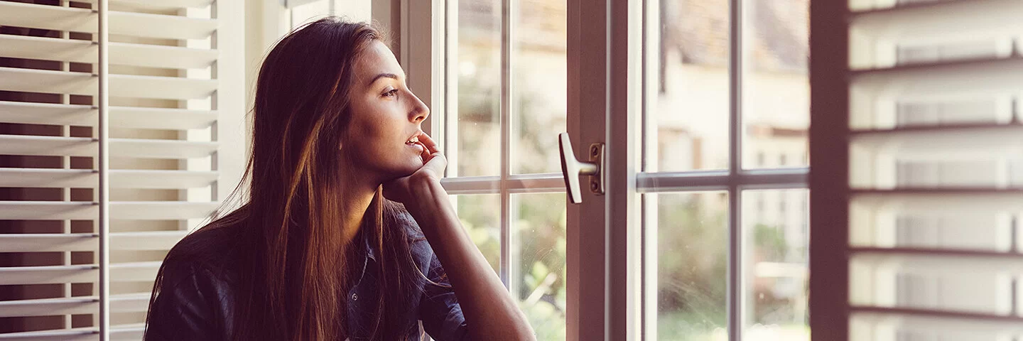 Eine junge Frau sitzt, das Kinn in eine Hand gestützt, an einem Fenster und schaut nach draußen.