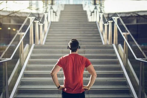 Ein Mann mit einem roten T-Shirt und Kopfhörern steht vor einer Treppe und blickt die Stufen herauf.
