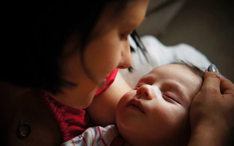 Nahaufnahme einer Mutter, die ihrem müden Säugling beim nächtlichen Stillen behutsam über die Stirn streicht, um es wach und trinkbereit zu halten.