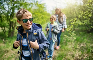 Eine erwachsene Frau und zwei etwa sieben und neun Jahre alte Jungs wandern im Frühling durch einen Wald. Alle tragen Jacken, lange Hosen und festes Schuhwerk zum Schutz vor Zecken.