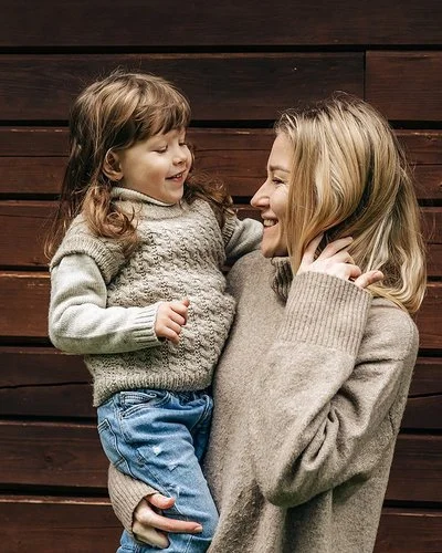 Eine lächelnde Frau hält ein lachendes kleines Mädchen auf dem Arm. Beide tragen Pullover aus Merinowolle in Naturfarben.