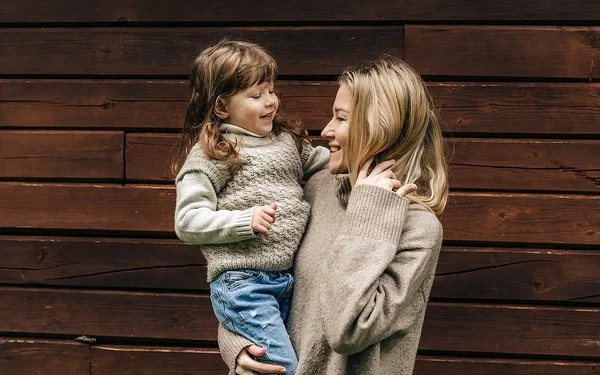 Eine lächelnde Frau hält ein lachendes kleines Mädchen auf dem Arm. Beide tragen Pullover aus Merinowolle in Naturfarben.