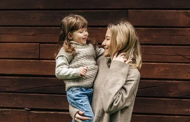 Eine lächelnde Frau hält ein lachendes kleines Mädchen auf dem Arm. Beide tragen Pullover aus Merinowolle in Naturfarben.