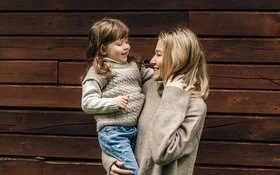 Eine lächelnde Frau hält ein lachendes kleines Mädchen auf dem Arm. Beide tragen Pullover aus Merinowolle in Naturfarben.