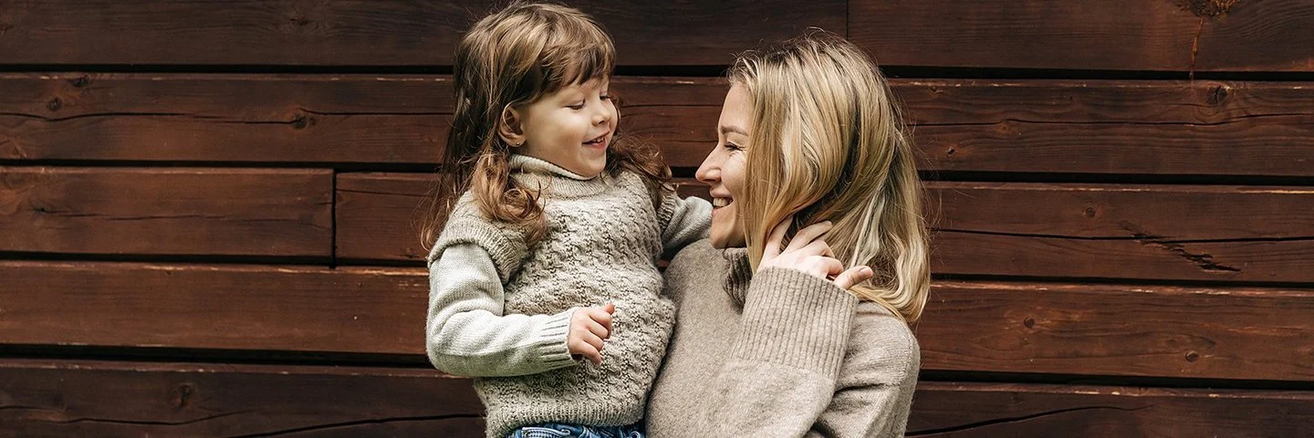 Eine lächelnde Frau hält ein lachendes kleines Mädchen auf dem Arm. Beide tragen Pullover aus Merinowolle in Naturfarben.