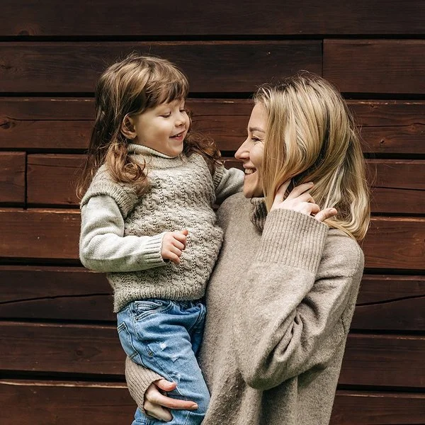 Eine lächelnde Frau hält ein lachendes kleines Mädchen auf dem Arm. Beide tragen Pullover aus Merinowolle in Naturfarben.