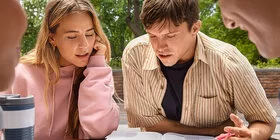 Zwei junge Frauen und zwei junge Männer sitzen im Freien an einem Tisch und lernen gemeinsam. Vor ihnen liegen verschiedene aufgeschlagene Bücher über die sie sich unterhalten. Im Hintergrund ist ein Baum zu sehen.