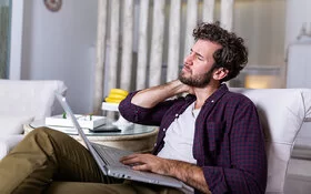 Ein junger Mann sitzt mit einem Laptop auf dem Schoß und geschlossenen Augen auf einem Sessel und fasst sich mit der rechten Hand an den Nacken.