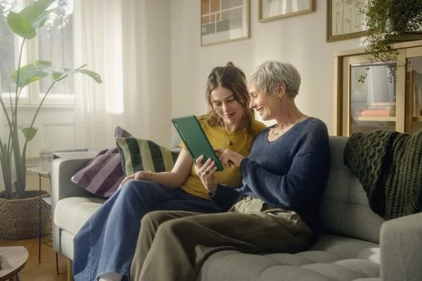 Eine ältere und eine jüngere Frau sitzen auf dem Sofa und schauen gemeinsam auf ein Tablet.