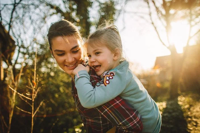 Mutter und Tochter halten lächelnd ihre Köpfe aneinander. Sie befinden sich in der Natur, das Licht ist sehr warm.