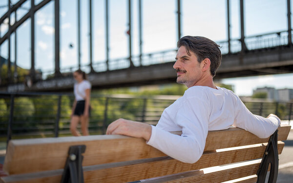 Arzt und Schauspieler Dr. Christoph-Gérard Stein sitzt auf einer Bank vor einer Brücke.