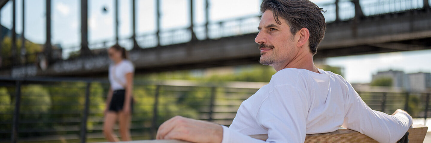 Arzt und Schauspieler Dr. Christoph-Gérard Stein sitzt auf einer Bank vor einer Brücke.