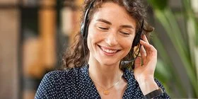 Eine junge Frau mit dunklem, lockigem Haar sitzt lächelnd mit einem Headset vor einem Laptop.