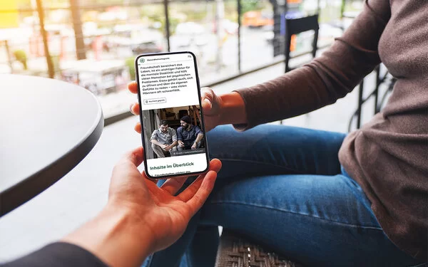 Eine Frau sitzt auf einer Terrasse und hält ein Smartphone in der Hand. Sie zeigt einer anderen Person den Bildschirm, auf dem ein Artikel aus dem AOK-Gesundheitsmagazin zu sehen ist.