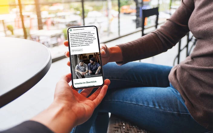 Eine Frau sitzt auf einer Terrasse und hält ein Smartphone in der Hand. Sie zeigt einer anderen Person den Bildschirm, auf dem ein Artikel aus dem AOK-Gesundheitsmagazin zu sehen ist.