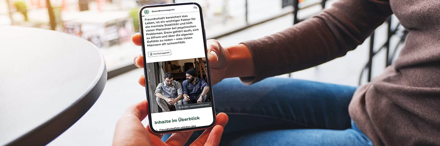 Eine Frau sitzt auf einer Terrasse und hält ein Smartphone in der Hand. Sie zeigt einer anderen Person den Bildschirm, auf dem ein Artikel aus dem AOK-Gesundheitsmagazin zu sehen ist.