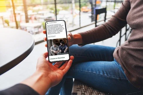 Eine Frau sitzt auf einer Terrasse und hält ein Smartphone in der Hand. Sie zeigt einer anderen Person den Bildschirm, auf dem ein Artikel aus dem AOK-Gesundheitsmagazin zu sehen ist.