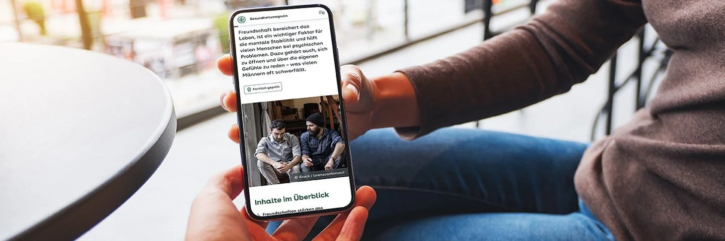 Nahaufnahme eines Smartphones, das von zwei Personen gehalten wird. Darauf ist ein Artikel mit dem AOK-Fachprüfungsstempel „Fachlich geprüft“ zu sehen.
