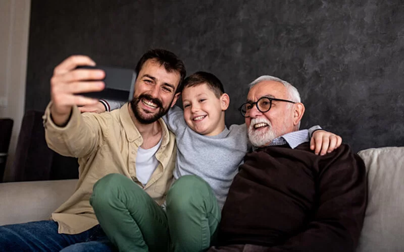 Ein Großvater, Vater und Enkel sitzen auf einem Sofa und machen ein Selfie.