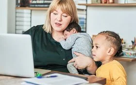 Eine Frau sitzt an einem Tisch im Wohnzimmer vor dem Computer. Auf ihrem Arm hält sie mit einer Hand ein schlafendes Baby, neben ihr sitzt ein Kleinkind und schaut ebenfalls auf den Bildschirm.