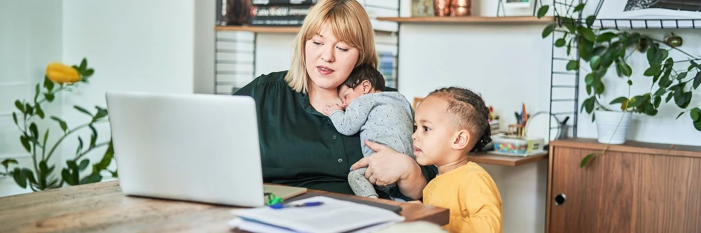 Eine Frau sitzt an einem Tisch im Wohnzimmer vor dem Computer. Auf ihrem Arm hält sie mit einer Hand ein schlafendes Baby, neben ihr sitzt ein Kleinkind und schaut ebenfalls auf den Bildschirm.