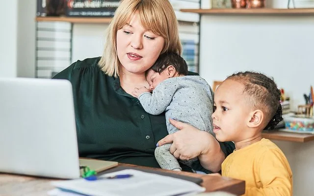 Eine Frau sitzt an einem Tisch im Wohnzimmer vor dem Computer. Auf ihrem Arm hält sie mit einer Hand ein schlafendes Baby, neben ihr sitzt ein Kleinkind und schaut ebenfalls auf den Bildschirm.