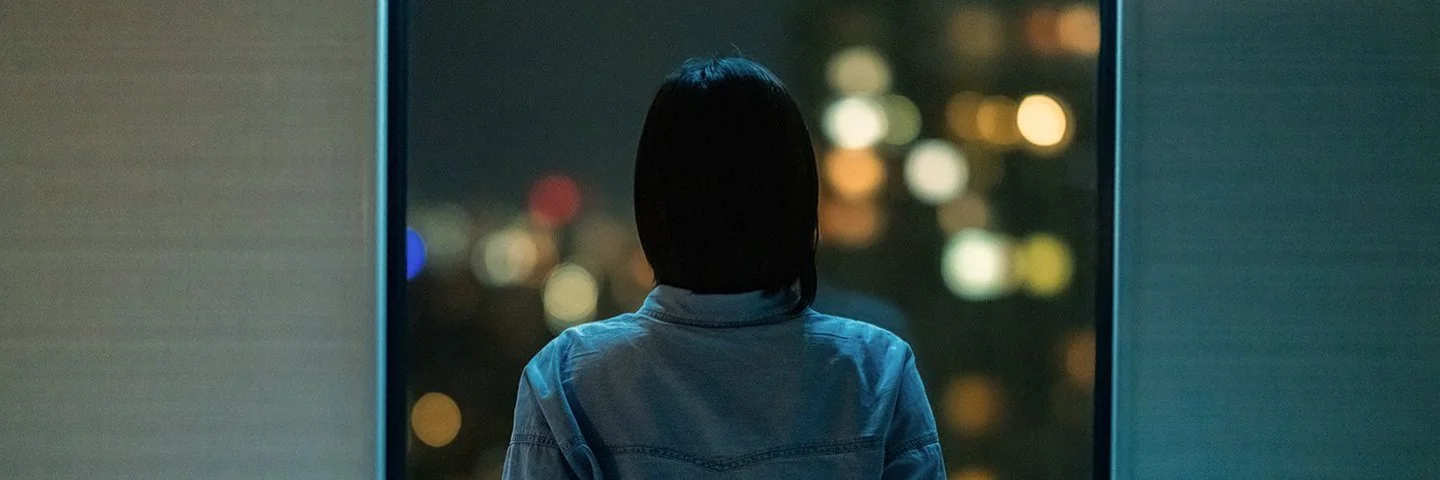 Eine Frau sitzt in einem dunklen Zimmer auf ihrem Bett und schaut aus dem Fenster. Draußen sind unscharfe Stadtlichter im Dunkeln zu sehen.