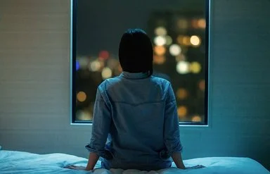 Eine Frau sitzt in einem dunklen Zimmer auf ihrem Bett und schaut aus dem Fenster. Draußen sind unscharfe Stadtlichter im Dunkeln zu sehen.