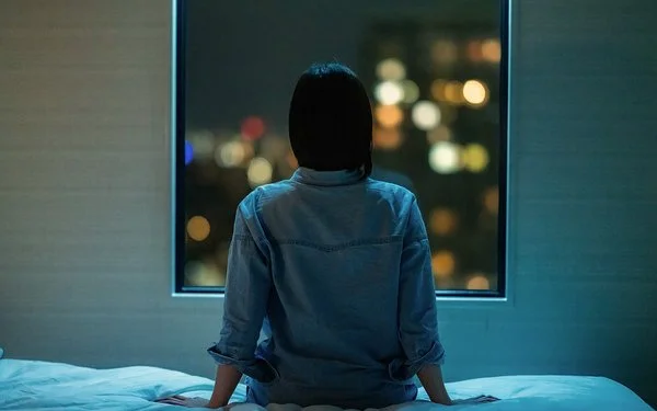 Eine Frau sitzt in einem dunklen Zimmer auf ihrem Bett und schaut aus dem Fenster. Draußen sind unscharfe Stadtlichter im Dunkeln zu sehen.