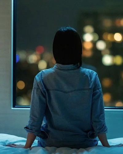 Eine Frau sitzt in einem dunklen Zimmer auf ihrem Bett und schaut aus dem Fenster. Draußen sind unscharfe Stadtlichter im Dunkeln zu sehen.