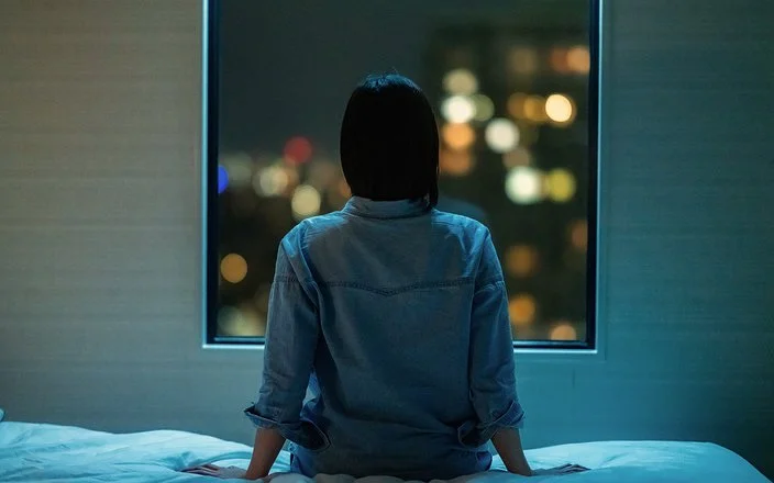 Eine Frau sitzt in einem dunklen Zimmer auf ihrem Bett und schaut aus dem Fenster. Draußen sind unscharfe Stadtlichter im Dunkeln zu sehen.
