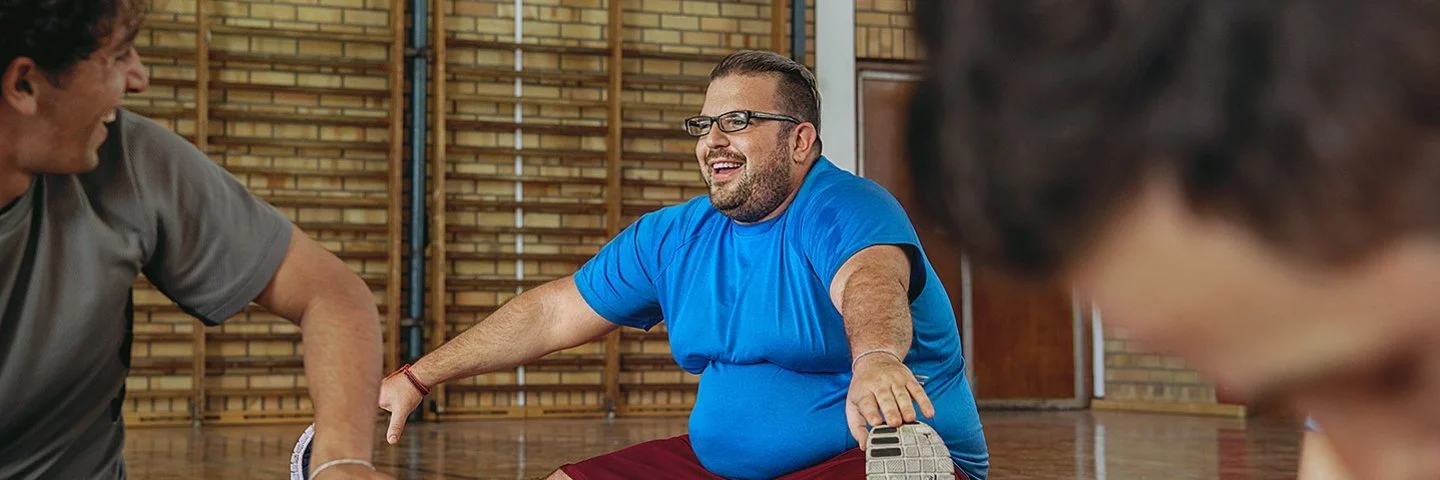 Ein Mann in blauem T-Shirt und roter Sporthose sitzt in einer Turnhalle beim Dehnen auf dem Boden, streckt ein Bein nach vorne und lächelt, während zwei weitere Personen unscharf im Vordergrund mittrainieren.